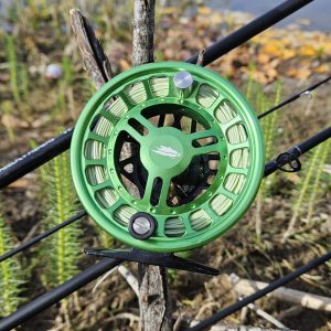 Reel Fly Fishers green chrono fly reel with versatile fly rod behind it, with a grassy beach behind them